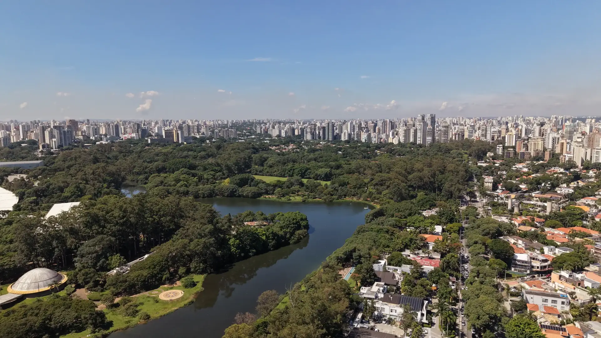 O lago do Parque Ibirapuera em curva, a mata, as casas do bairro e a cidade ao fundo. Foto aérea.
